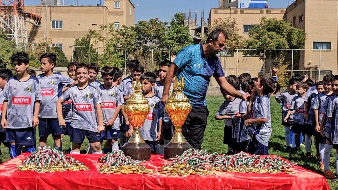 بازیکنان فوتبال نونهالان خوشحال با لباسهای طوسی و سرمه ای درزمین چمن درحال گرفتن مدال و جام قهرمانی از مربی خود